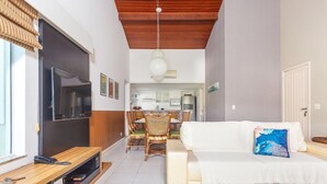 A living room with a TV, wooden TV stand, white sofa, dining table with chairs, and a kitchen in the background. - Bz73 Geribá! Casa c/ piscina privativa 5 Suítes! (Búzios)