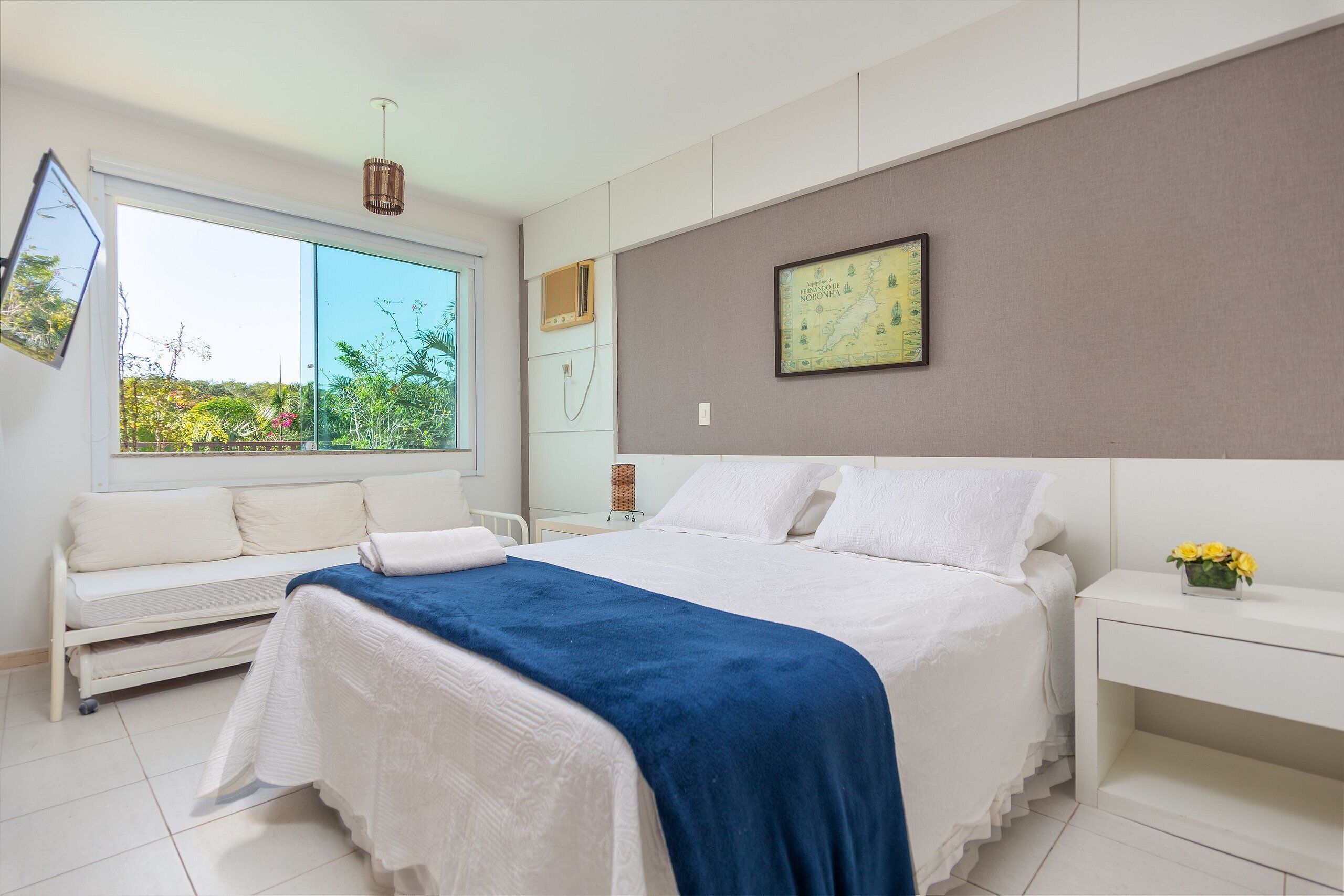 A modern bedroom with a large bed, a bedside table with a vase of flowers, a map on the wall, and a window with a view of greenery.