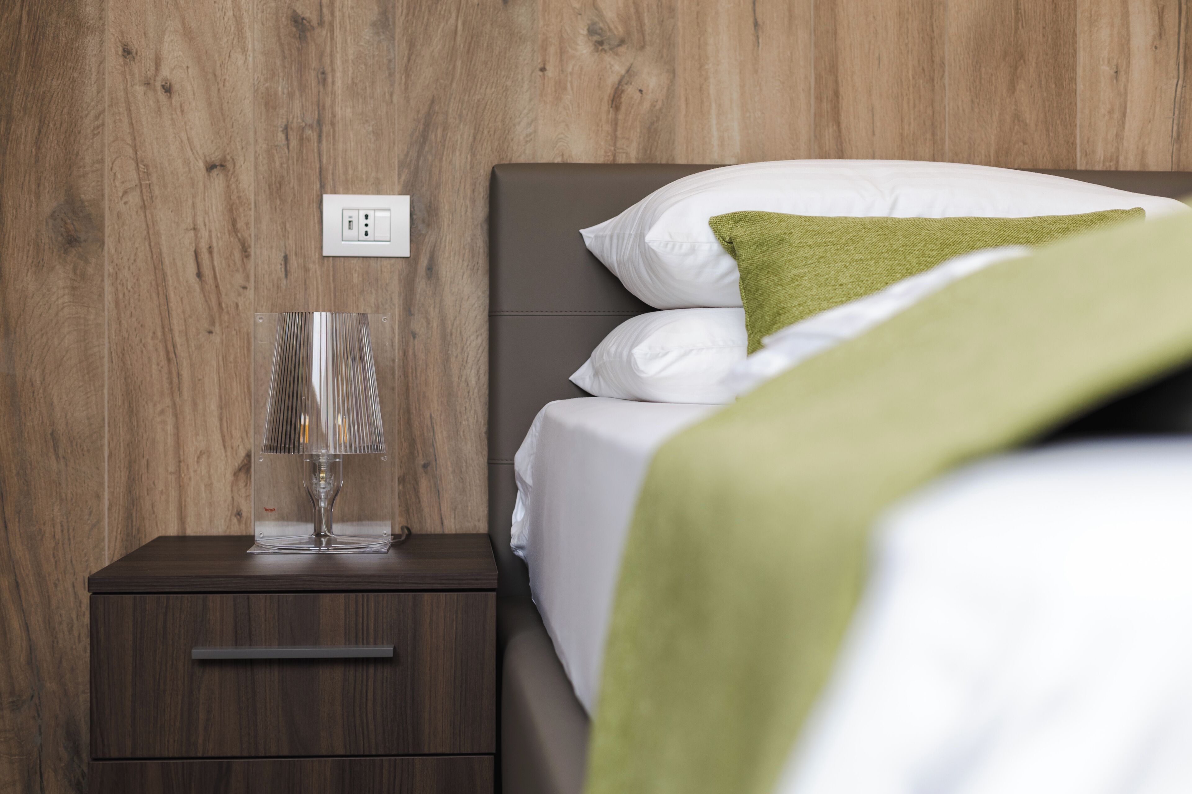 A neatly made bed with white and green bedding, a bedside lamp, and a wooden nightstand with a drawer.