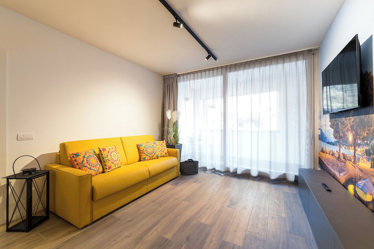 Ein modernes Wohnzimmer mit einem gelben Sofa, einem flachbildfernseher an der Wand und einem großen Fenster mit durchsichtigen Vorhängen.