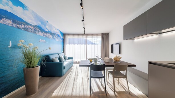 A modern living room with a large mural of a coastal scene, a blue sofa, a dining table with chairs, and a kitchen area with a refrigerator and cabinets. - Boutique Apartments By Annalisa (Nago-Torbole)