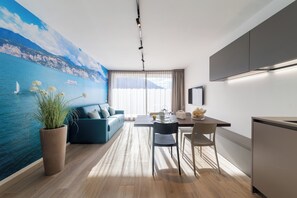 Una sala moderna con un gran mural de paisaje costero, un sofá azul, una mesa de comedor con sillas y una zona de cocina con refrigerador y armarios.