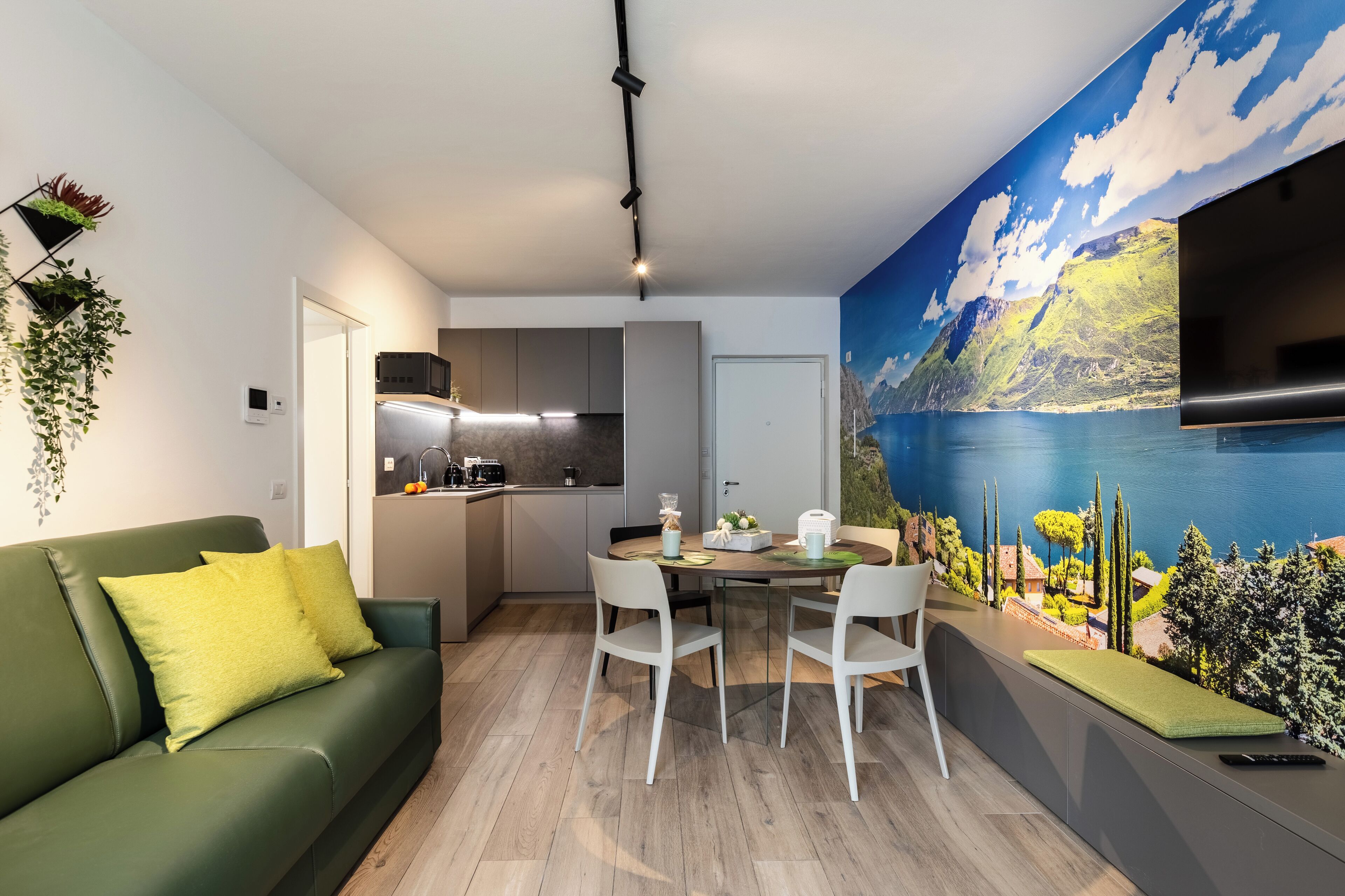 Ein modernes Wohnzimmer mit einem grünen Sofa, weißen Sesseln und einem Esstisch. Im Hintergrund ist eine Küche mit einem großformatigen Wandbild von einer Berglandschaft zu sehen.