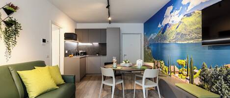 A modern living room with a green sofa, white chairs, and a dining table. There is a kitchen in the background with a large mural of a mountainous landscape.