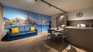 A modern living room with a blue sofa, dining table, and chairs. A kitchen area with a counter and appliances is visible. A large window offers a scenic view of a coastal town. - Boutique Apartments By Annalisa (Nago-Torbole)