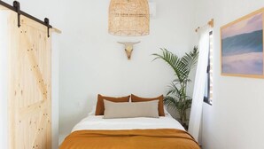 Une chambre à coucher avec un lit, une porte en bois, un luminaire fixé au mur, une plante et un tableau.