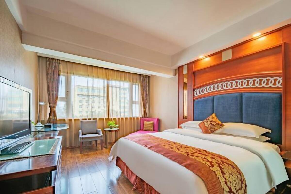 A hotel room with a bed, a desk with a chair, a TV, and a window with curtains.