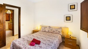 Quarto com cama, duas mesas de cabeceira, um criado-mudo, uma porta e luminária fixada na parede.