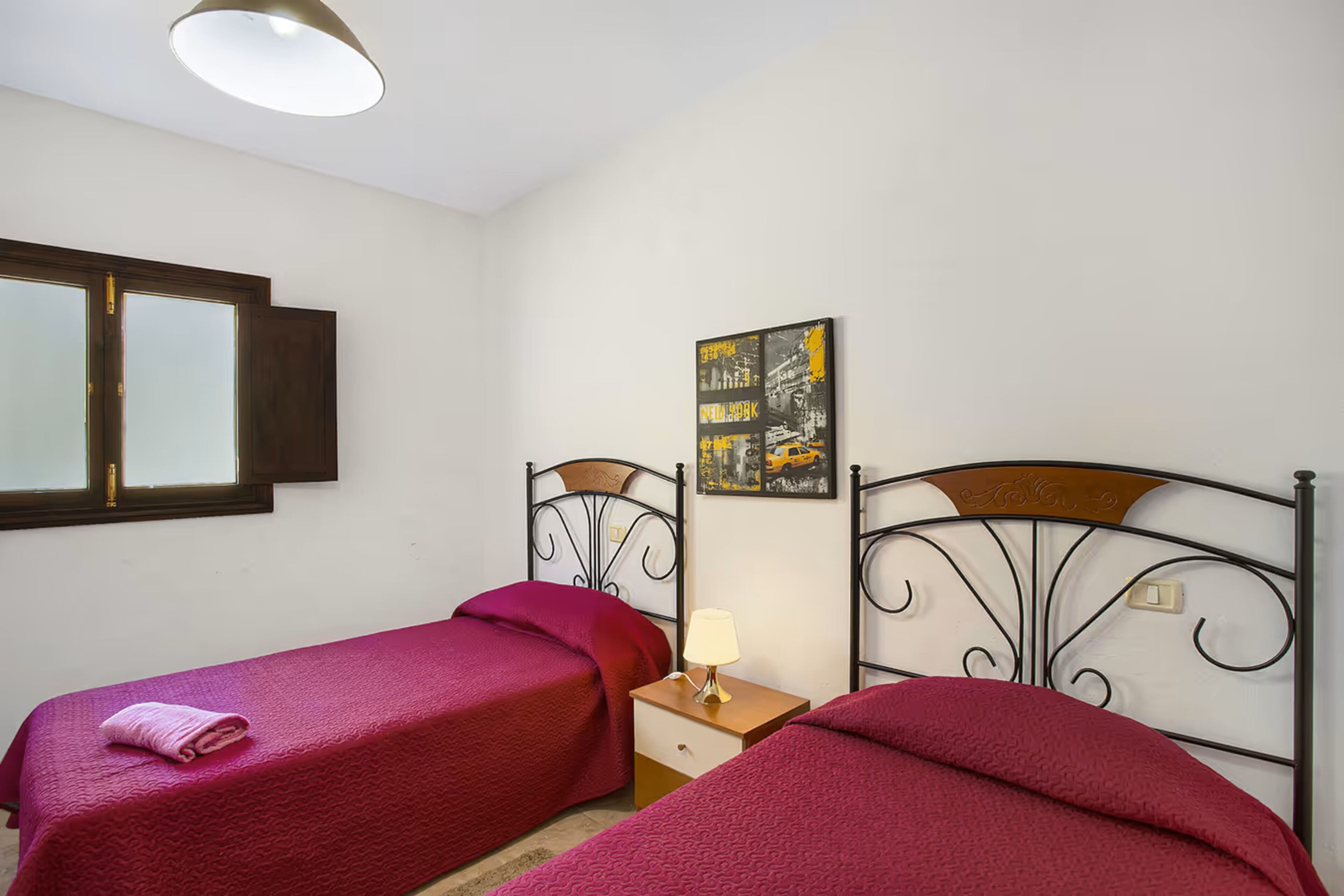 A bedroom with two single beds, a nightstand with a lamp, a window with blinds, and a wall-mounted artwork.