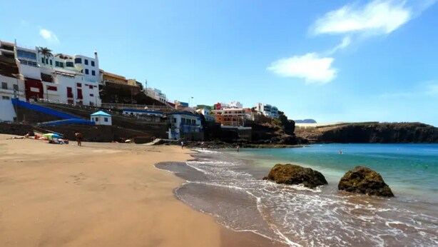 A sandy beach with clear blue water, rocky outcrops, and buildings in the background. - House Altos de Gáldar (Galdar)