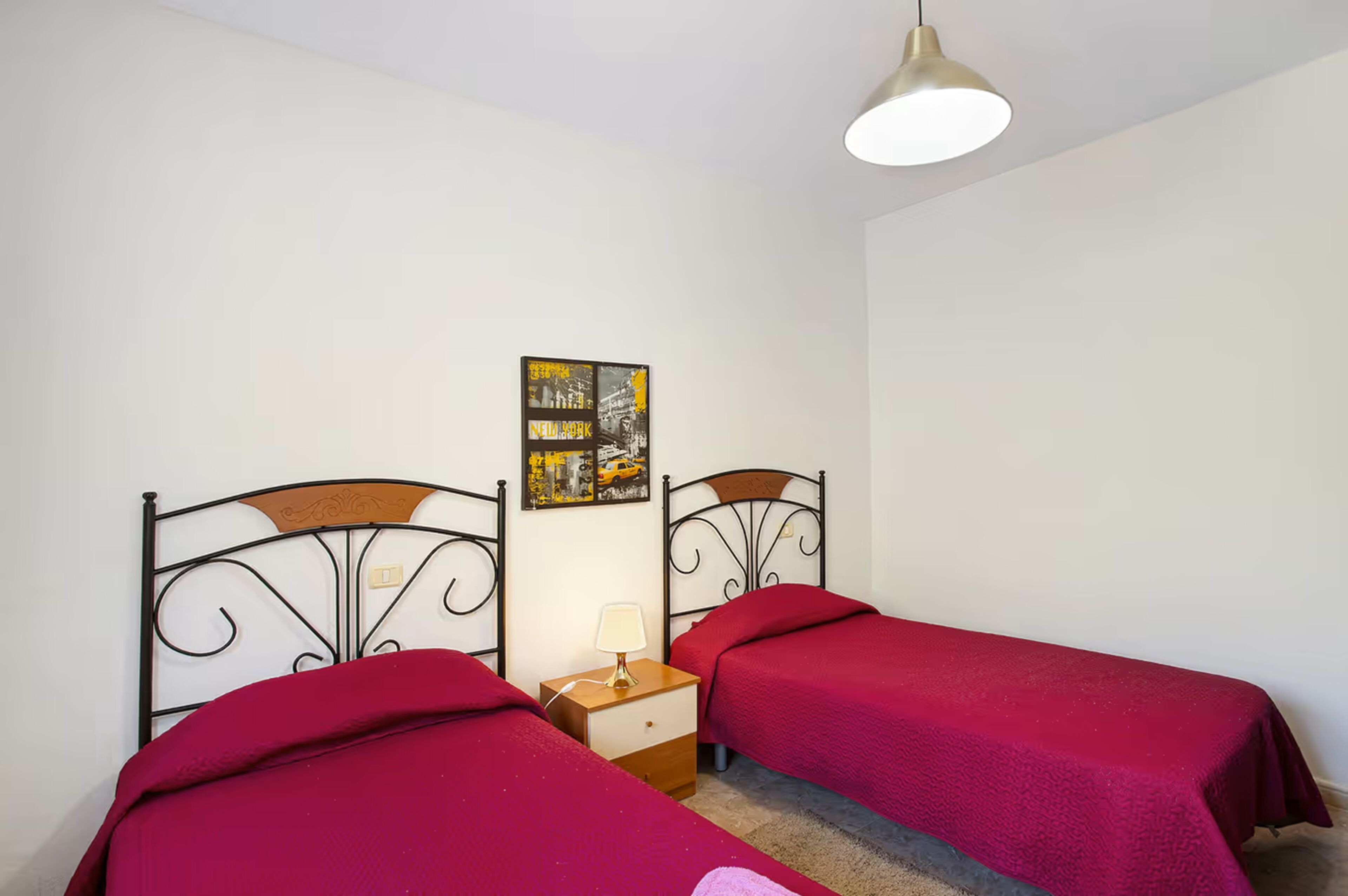 Une chambre avec deux lits, chacun recouvert d’une couverture rouge, une table de chevet avec une lampe et un tableau accroché au mur.