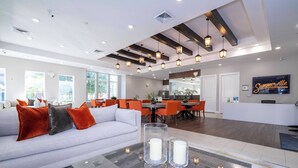 A modern hotel lobby with a light gray sofa, orange cushions, a glass-top coffee table with candles, and a large screen displaying 'Summerville'.