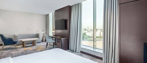 Une chambre d’hôtel avec un grand lit, un bureau, une chaise, un canapé, une télévision et de grandes fenêtres équipées de rideaux.