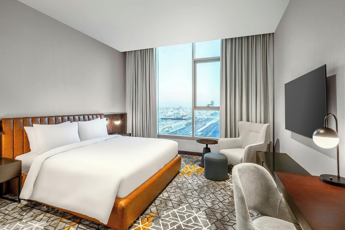 Hotelkamer met een groot bed, een bureau, een stoel, een televisie en uitzicht op de stad door een groot raam.