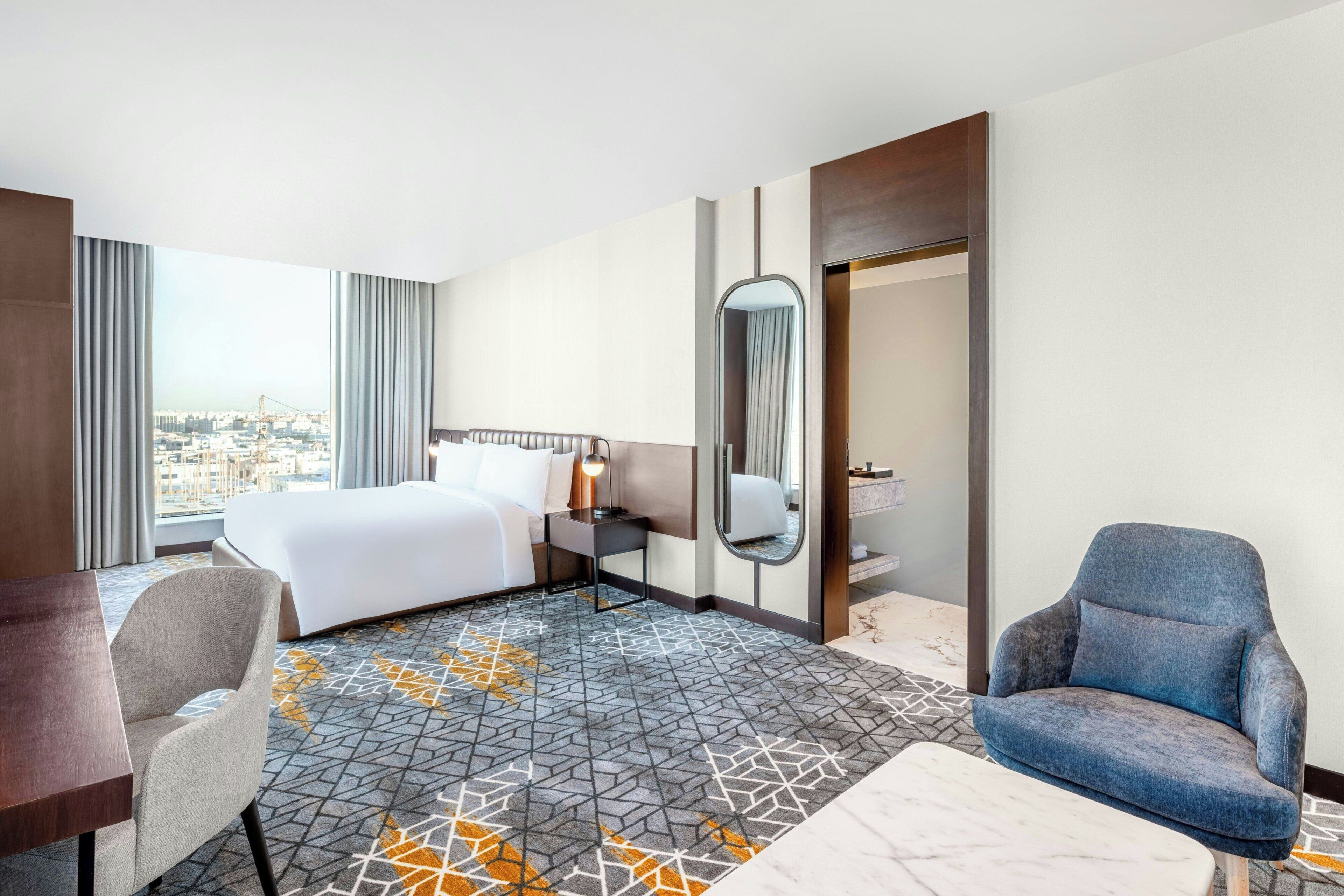 Une chambre d’hôtel avec un lit, un bureau, une chaise, un grand miroir et une vue sur la ville.