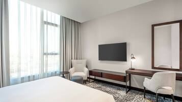Habitación de hotel con una cama grande, un escritorio con silla, un televisor montado en la pared y una ventana con cortinas.