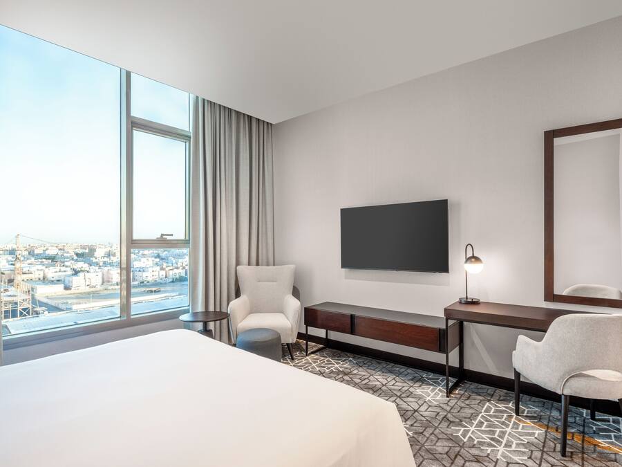 A hotel room with a large bed, a desk with a chair, a TV, and a view of the city through large windows.