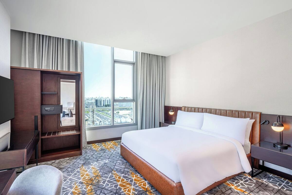 Een hotelkamer met een groot bed, een bureau met stoel, een televisie, een kledingkast en uitzicht op de stad door grote ramen.