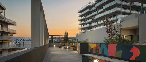 A rooftop terrace with a view of the sunset, a seating area with a colorful mural, and a building with multiple balconies.