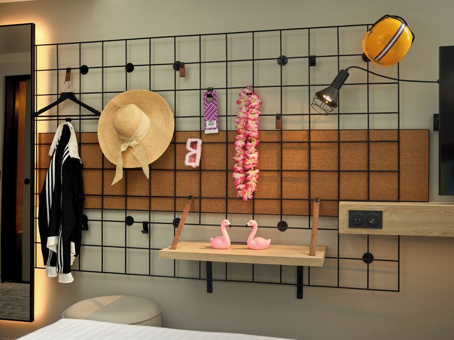 A modern hotel room with a bed, a mirror, a wall-mounted shelf with decorative items, and a corkboard.