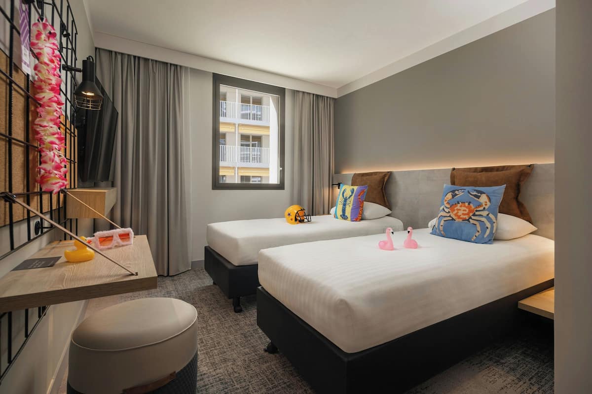 A hotel room with two beds, a desk, a chair, a window with curtains, and a view of a building.