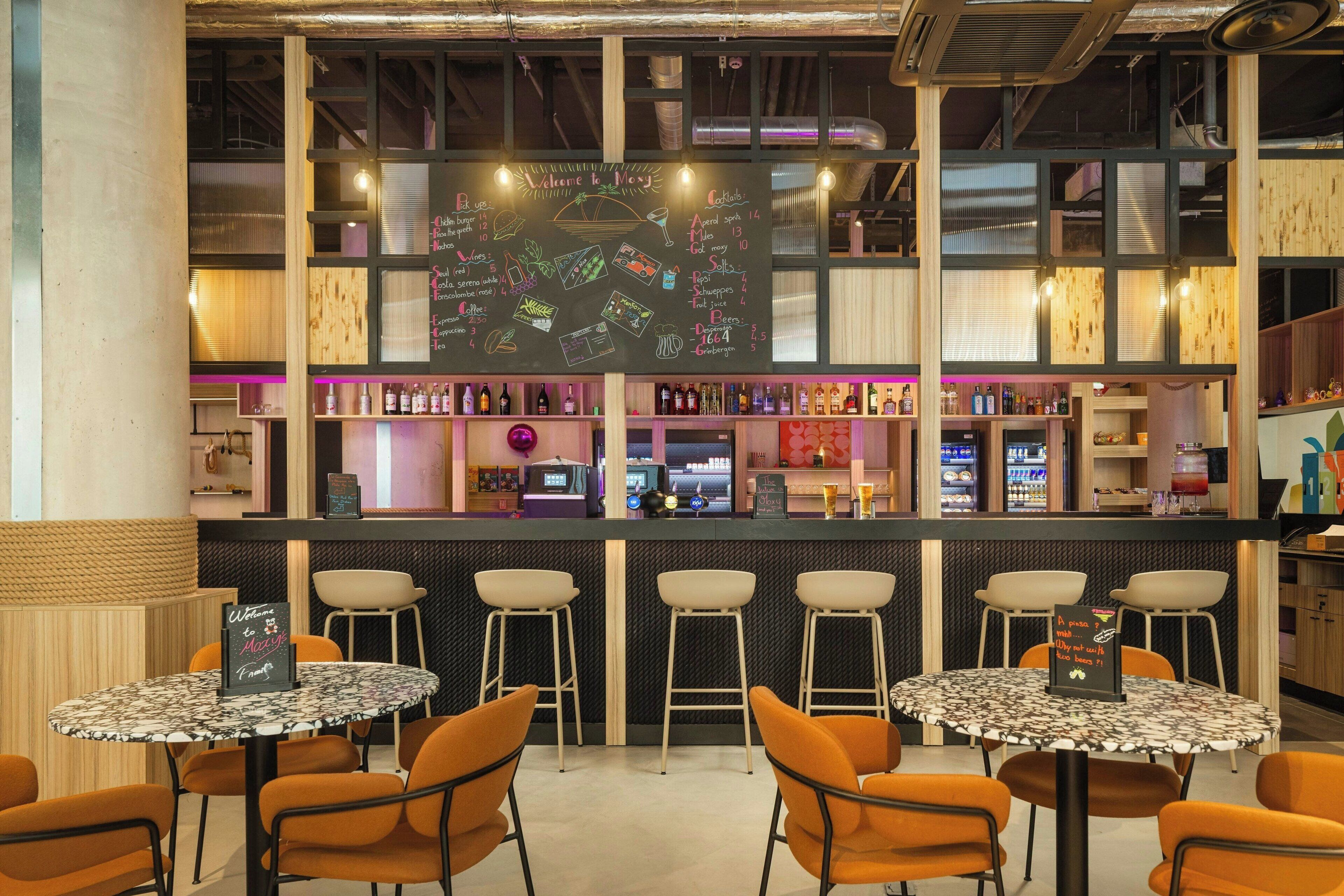 A bar area with a chalkboard menu, bar stools, and a seating area with round tables and orange chairs.