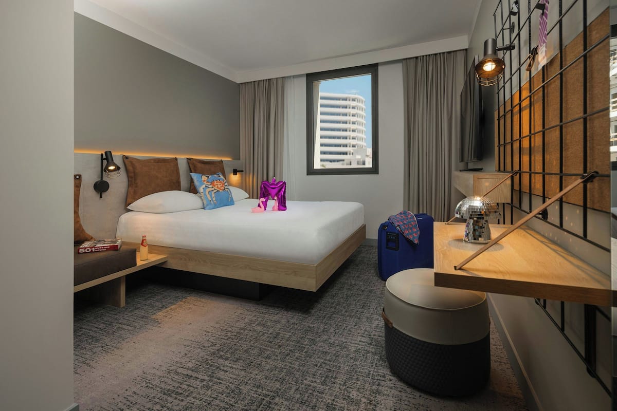 A modern hotel room with a large bed, a desk with a lamp, a suitcase, and a view of a building through the window.