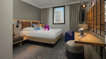 Habitación de hotel moderna con una cama grande, un escritorio con lámpara, una maleta y vistas a un edificio a través de la ventana.