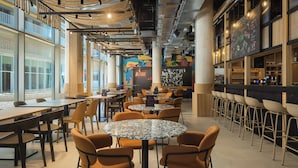 Una cafetería moderna con mesas redondas y sillas anaranjadas, ventanas amplias y un mural colorido en la pared.