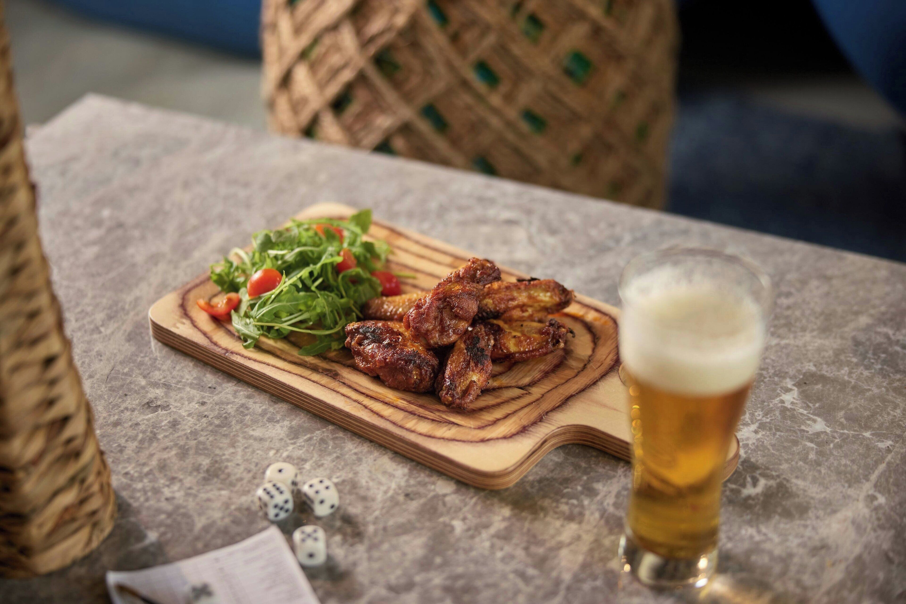 A meal of grilled chicken wings with a side salad and a glass of beer on a wooden board.