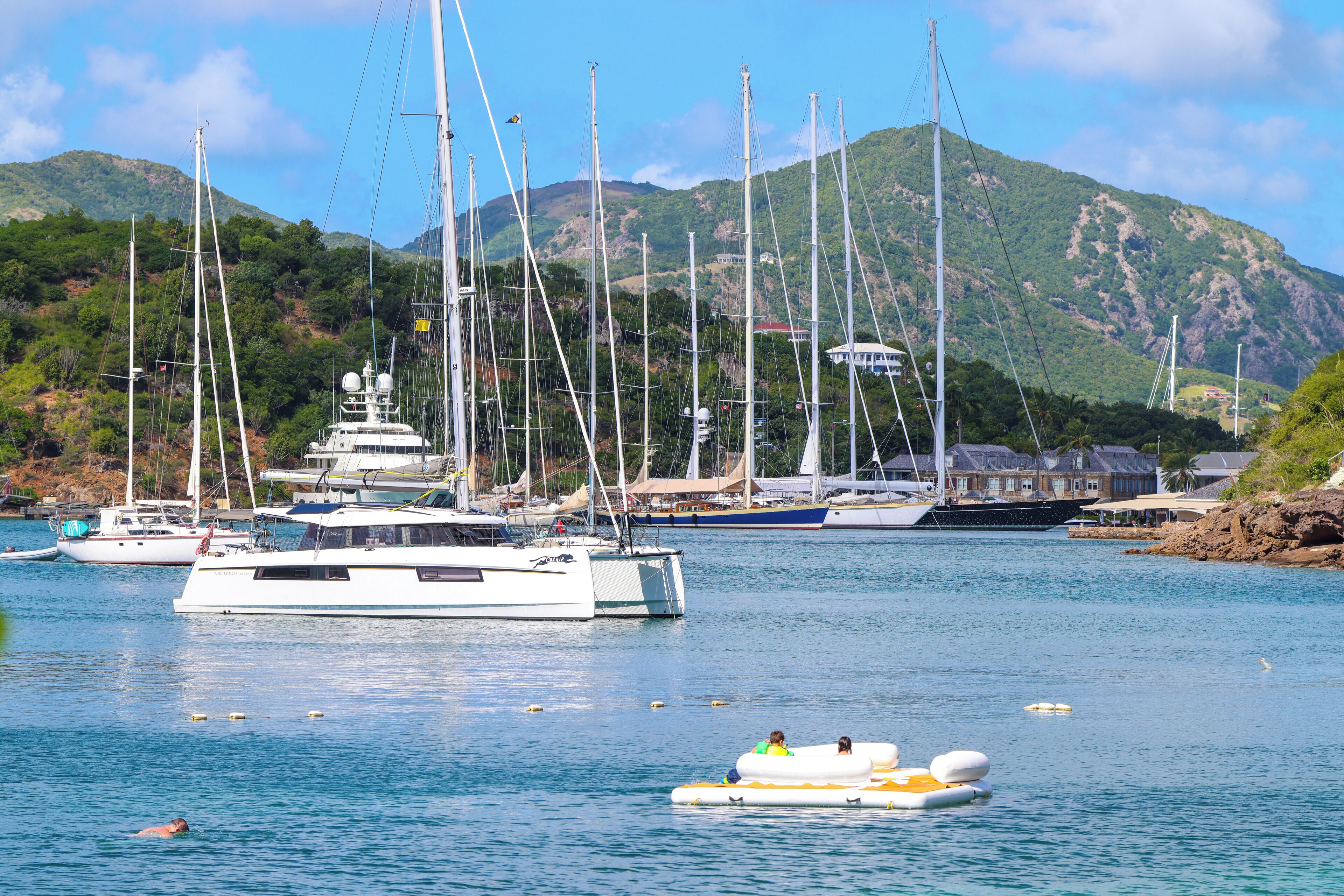 A marina with multiple sailboats and yachts docked, surrounded by lush green hills and clear blue water.