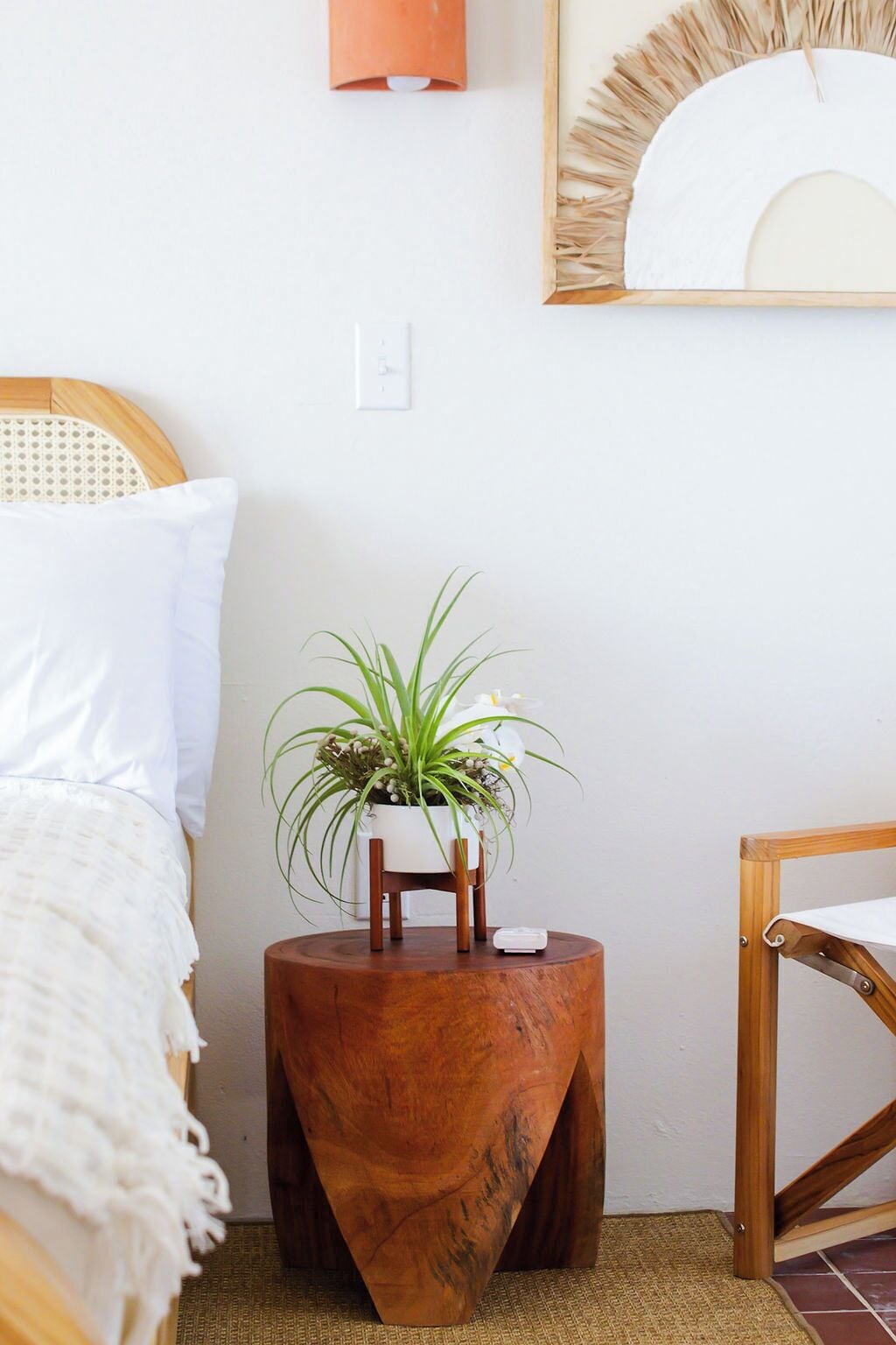 A bedroom with a bed, a bedside table with a plant, a wooden chair, and a round wooden stool.