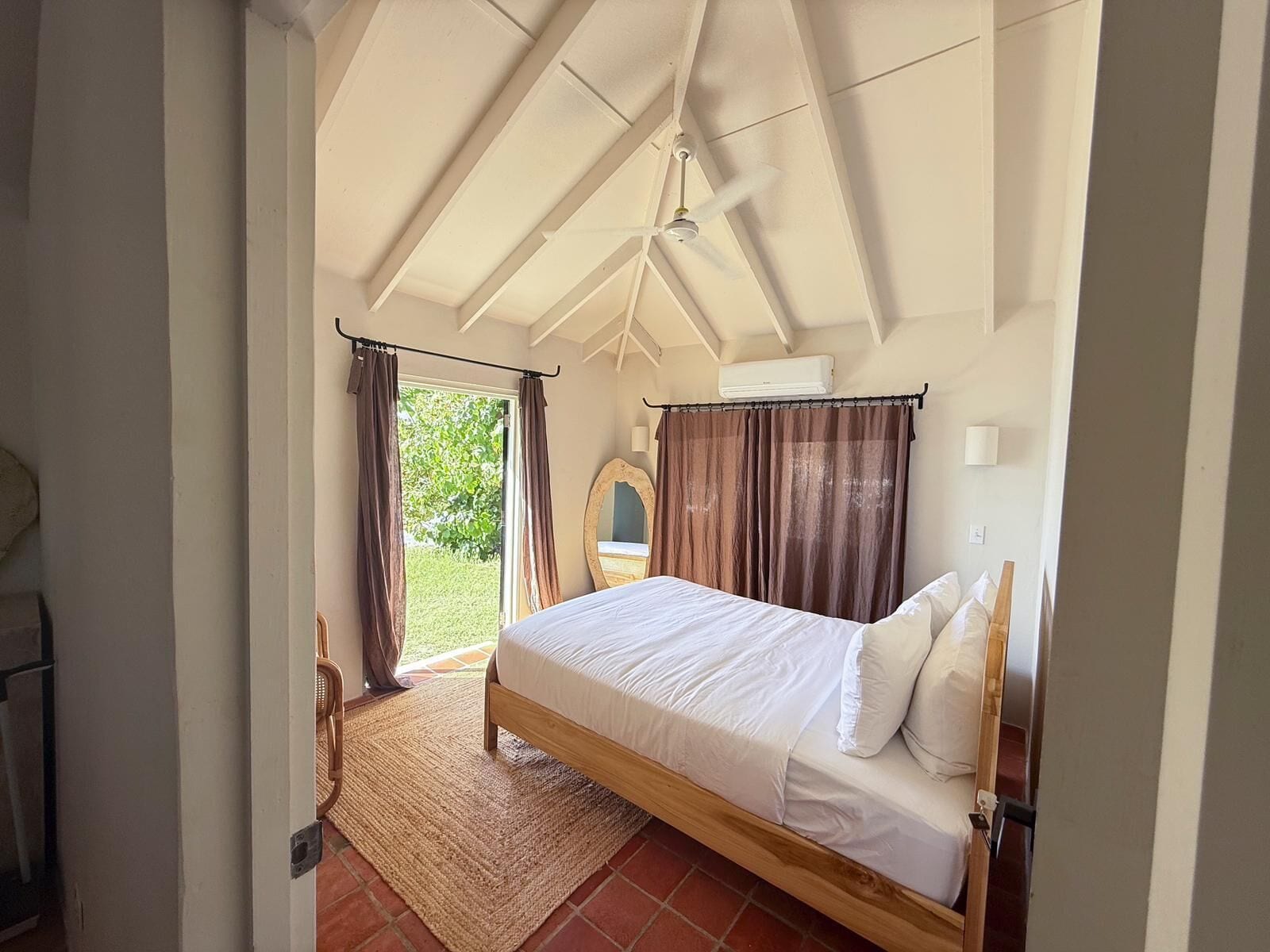 Un dormitorio con cama, ventilador de techo, ventana con cortinas y vista a zonas verdes.