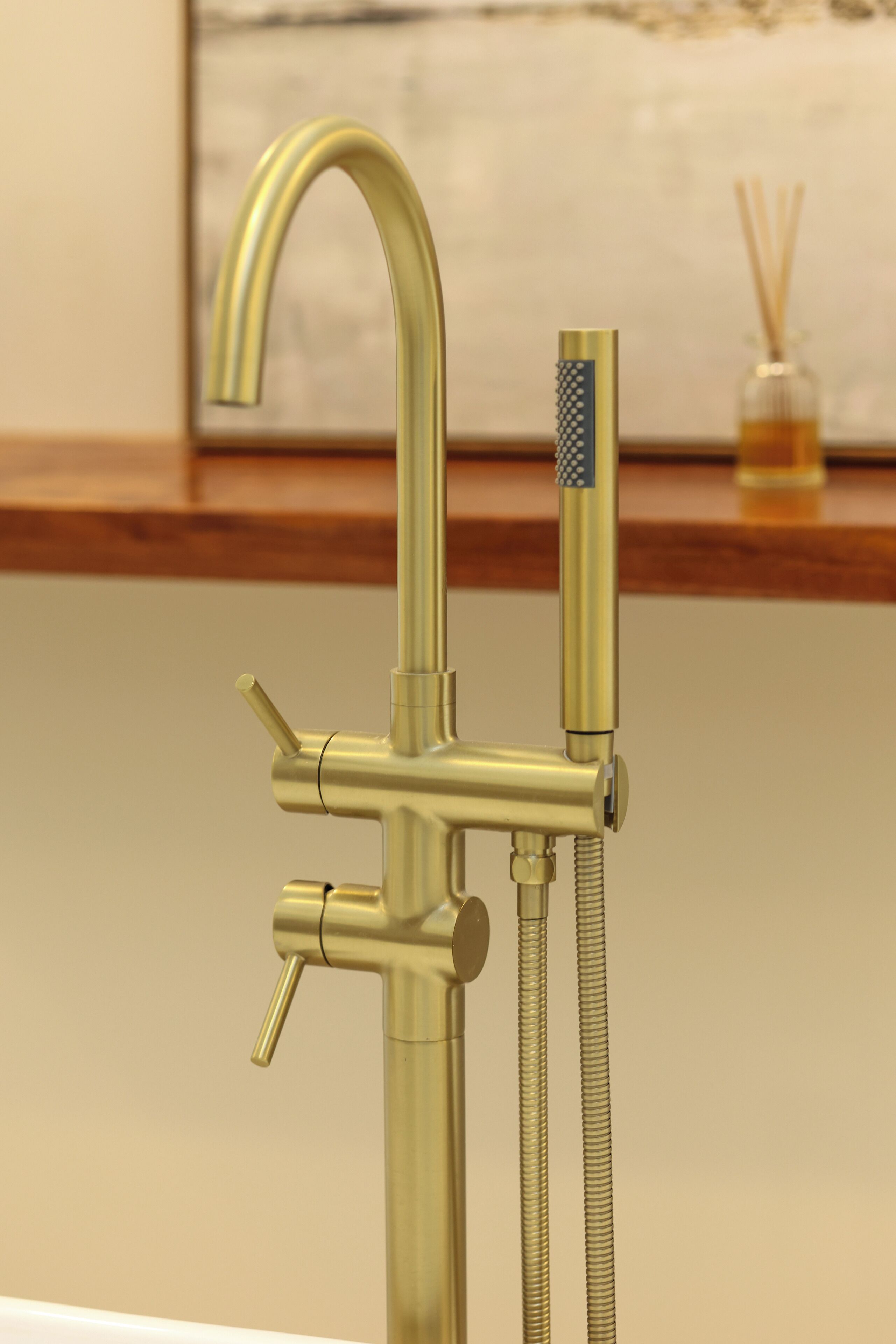 Une salle de bain moderne avec un robinet et une pomme de douche de couleur dorée, une douchette à main et un récipient en verre contenant des diffuseurs à bâtonnets, le tout posé sur une étagère en bois.