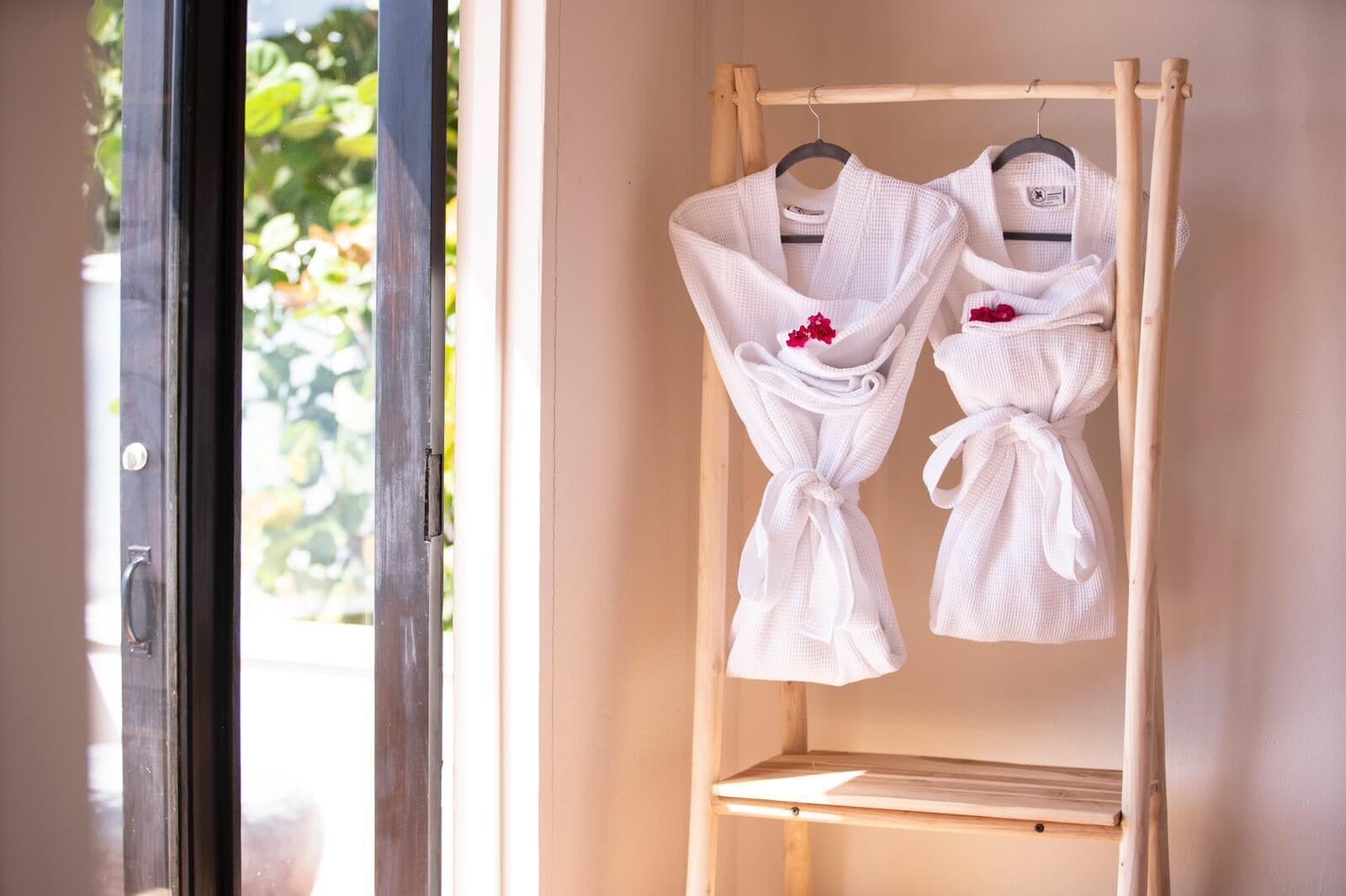 A wooden rack with two white robes tied at the waist, hanging on a wall near a window.