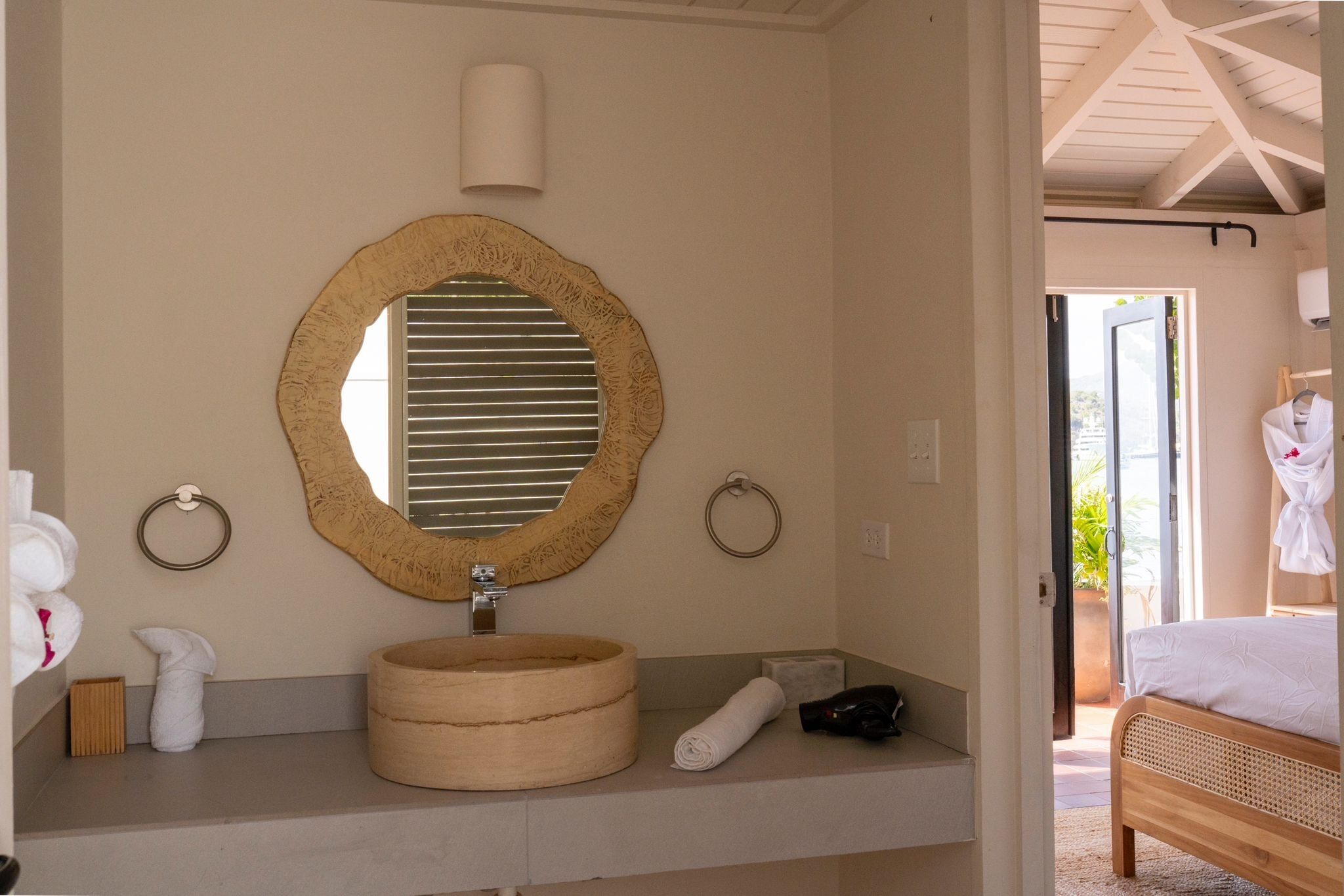 A bathroom with a round mirror, wooden sink, towel rings, and a bed visible through an open door.