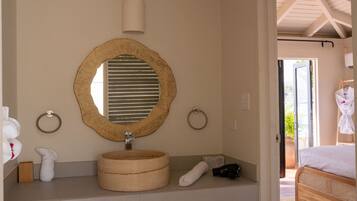 A bathroom with a round mirror, wooden sink, towel rings, and a bed visible through an open door.