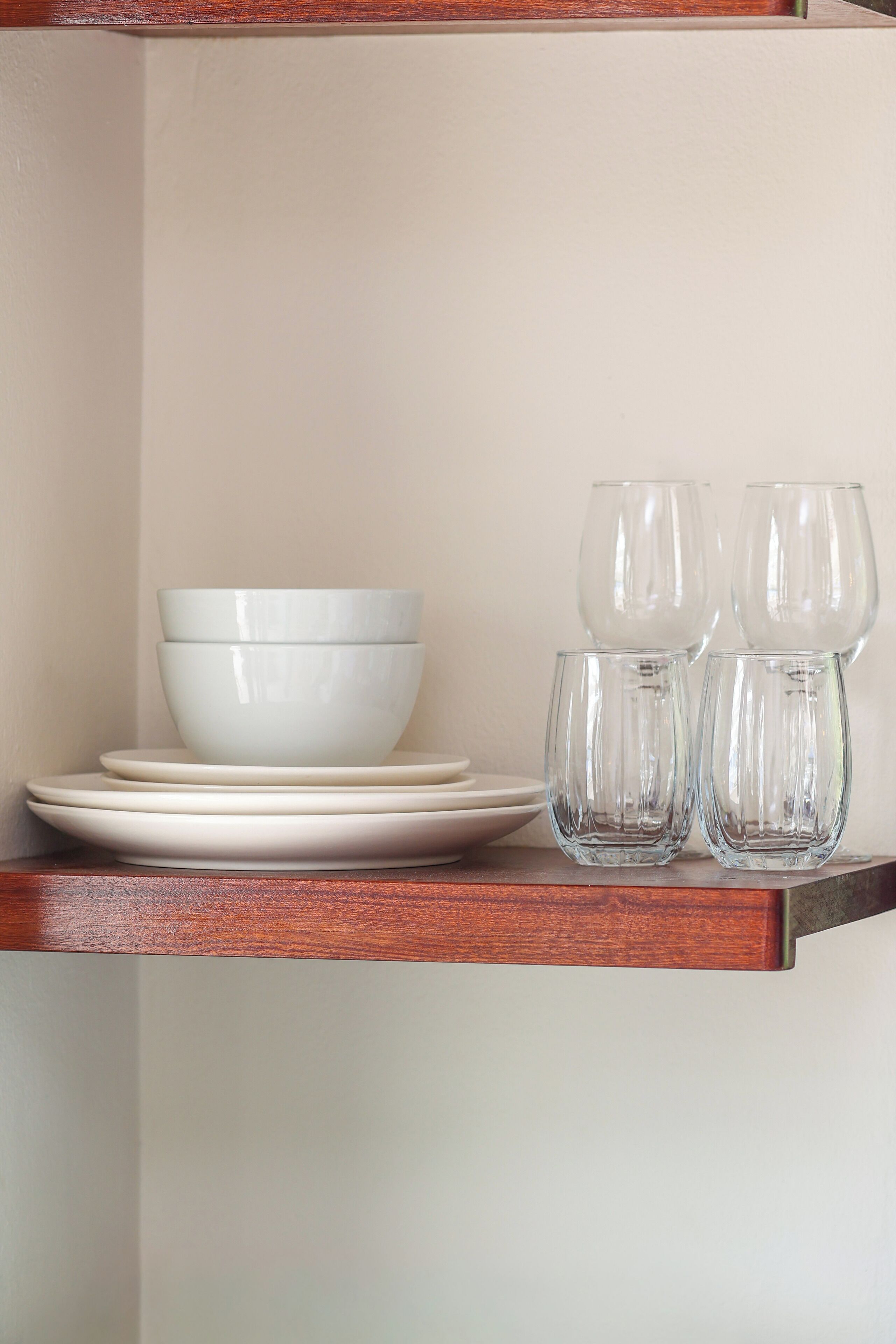 A set of white bowls and glasses on a wooden shelf against a plain wall.