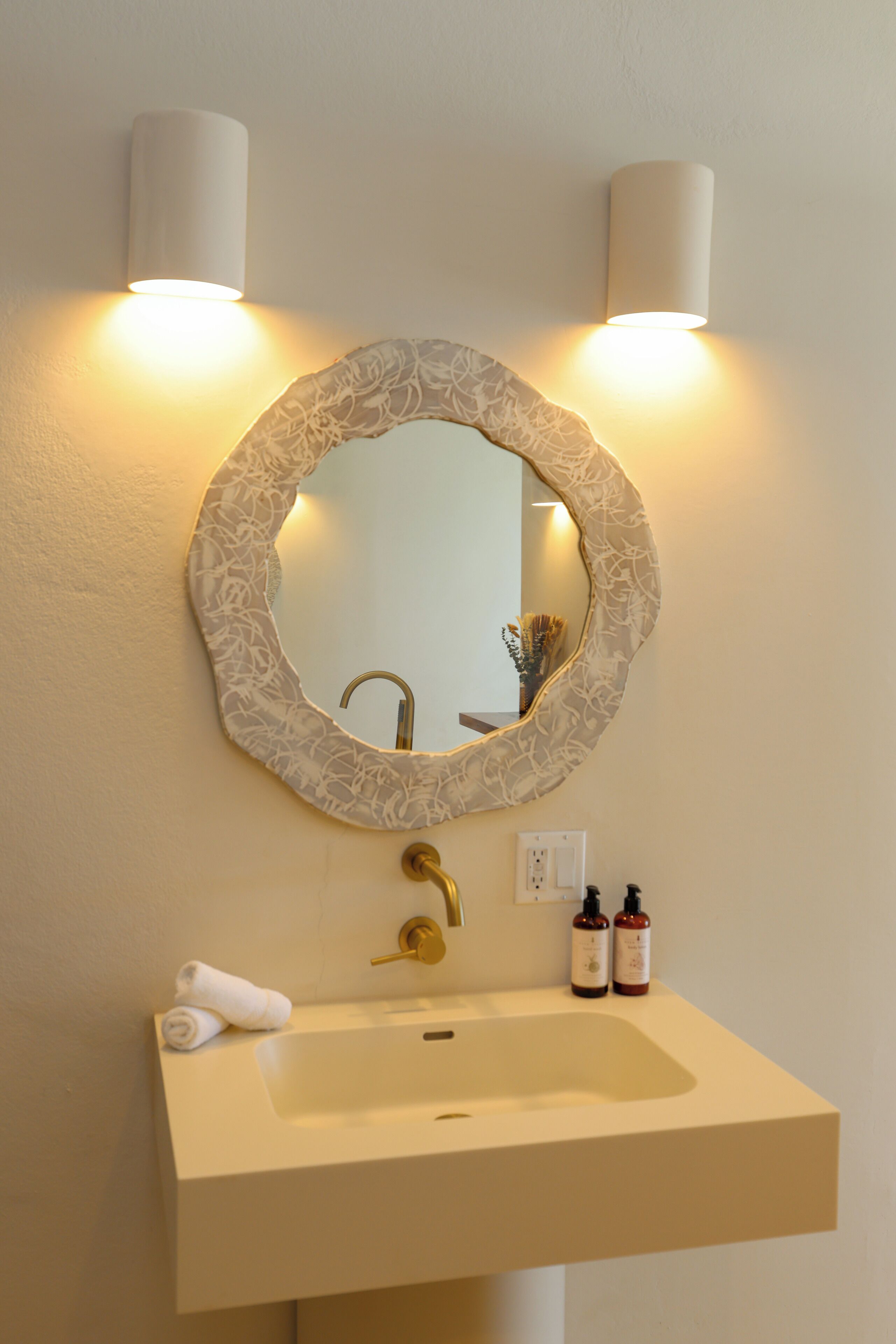 A bathroom with a round mirror, two wall-mounted lights, a sink, and two bottles of toiletries.