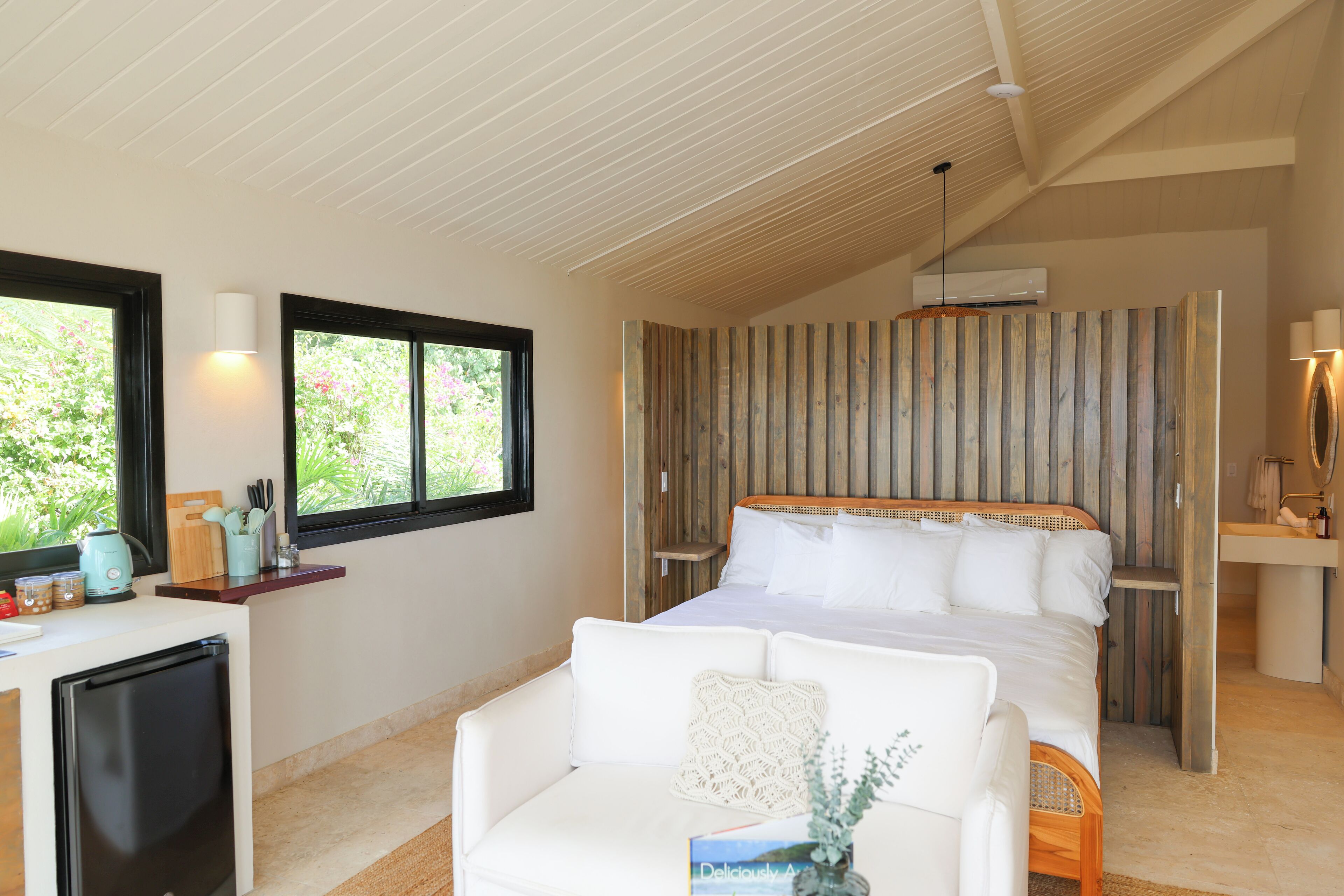 A bedroom with a bed, a white sofa, a desk with a computer, and a window with a view of greenery.