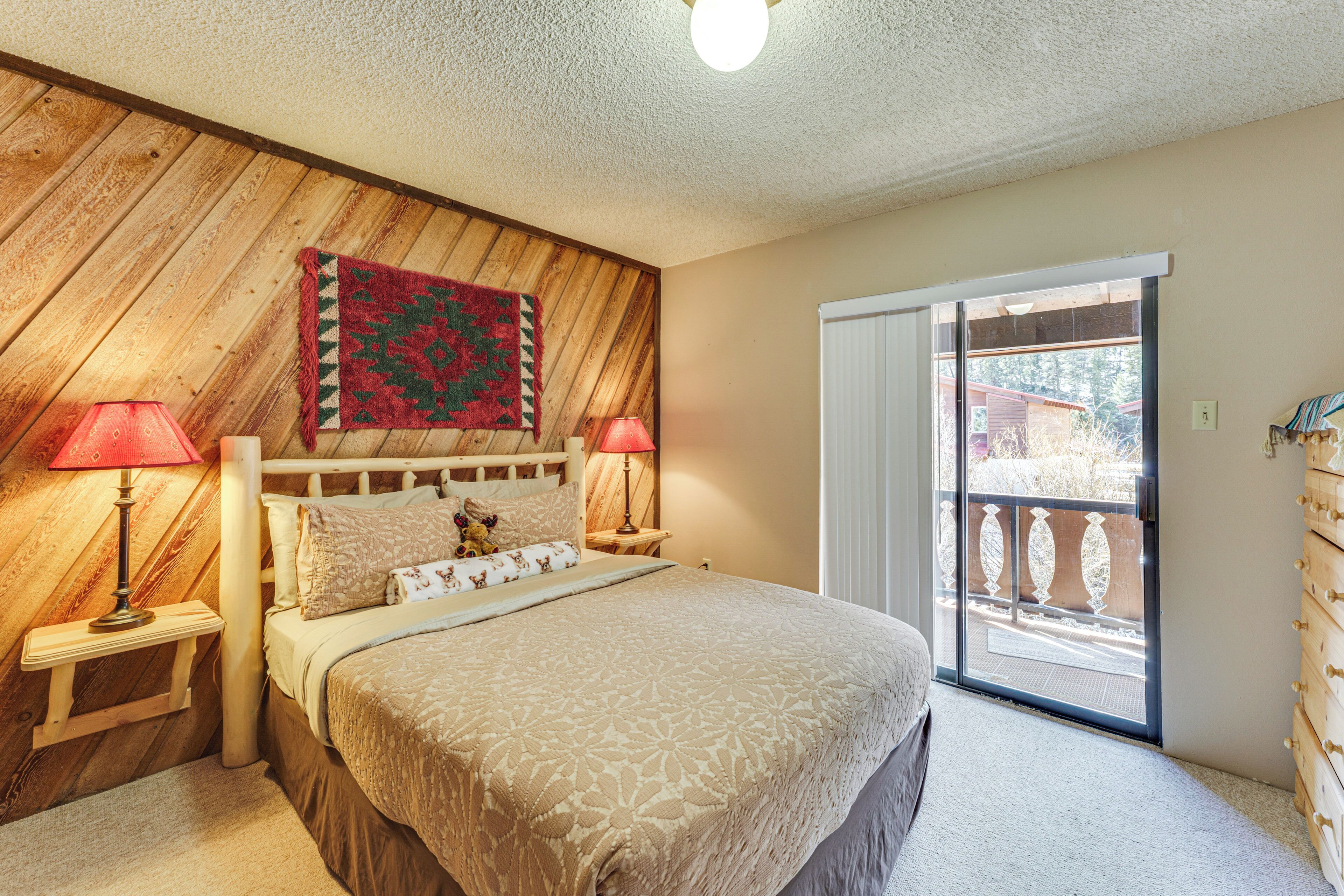 A bedroom with a wooden headboard, a large bed with patterned bedding, two bedside lamps, a red and green wall hanging, and a sliding glass door to the outside.