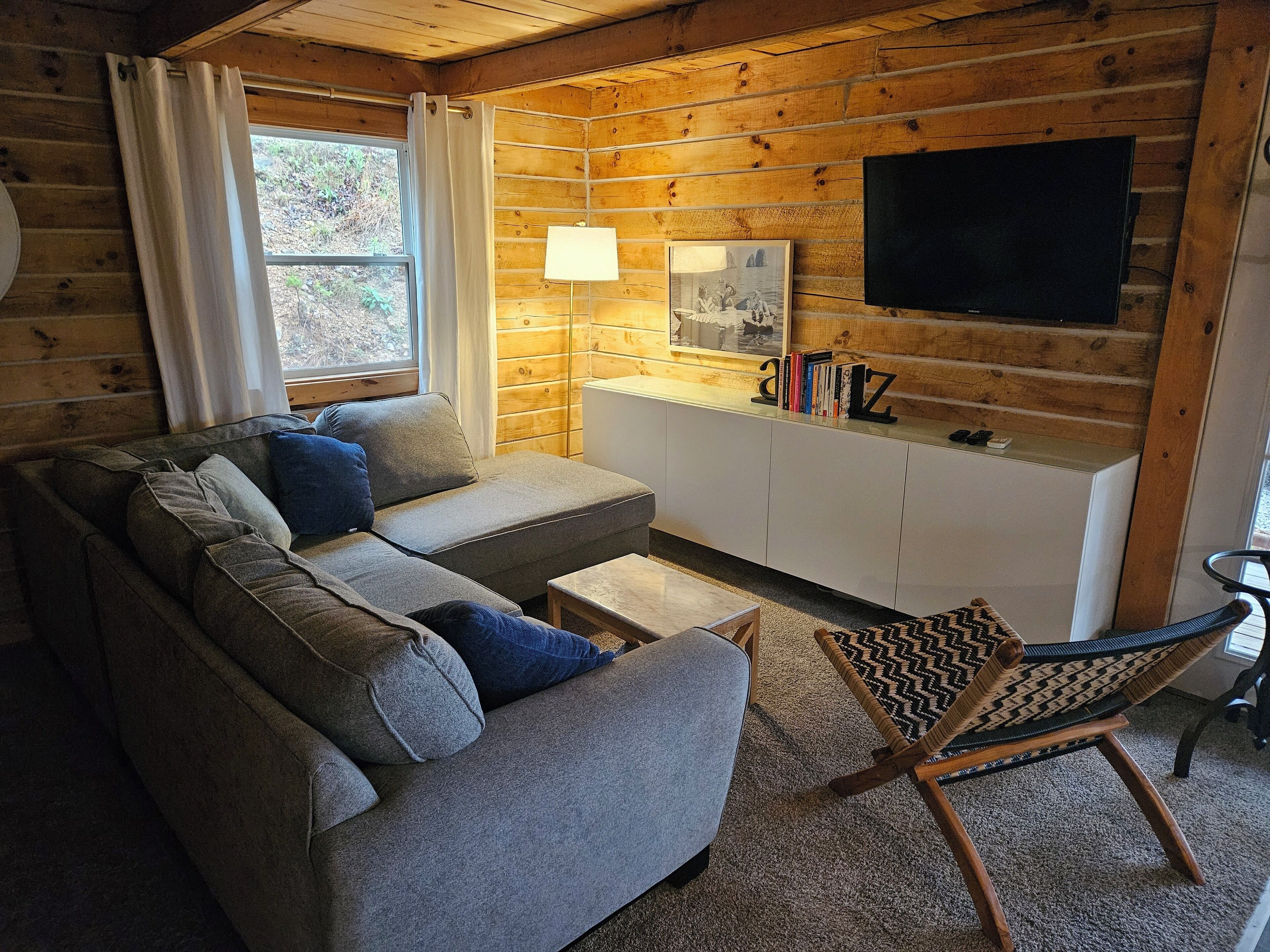 A cozy cabin-style room with a gray sofa, a small table, a patterned chair, a TV, and a bookshelf with books and a lamp.