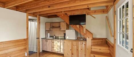 Une petite cabane en bois comprenant un coin cuisine, un petit réfrigérateur et un escalier menant à une porte.