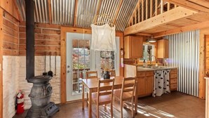 A rustic cabin interior with a wooden dining table, chairs, a kitchen area with a sink and cabinets, and a wood-burning stove.