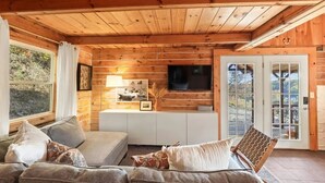 A cozy cabin interior with a wooden ceiling, a sofa, a TV, and a large window.
