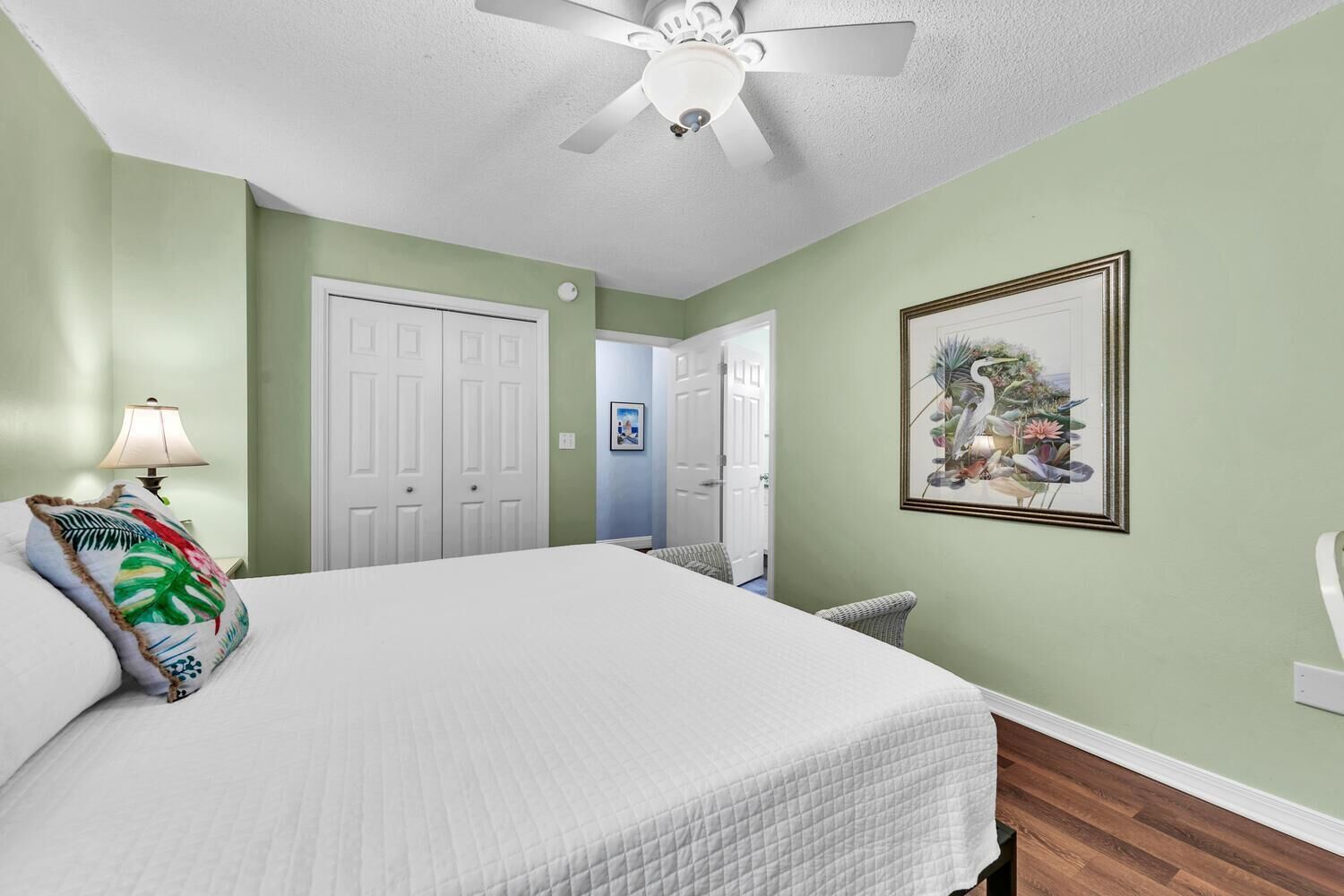 A bedroom with a bed, a ceiling fan, a nightstand with a lamp, a framed picture on the wall, and a door leading to another room.