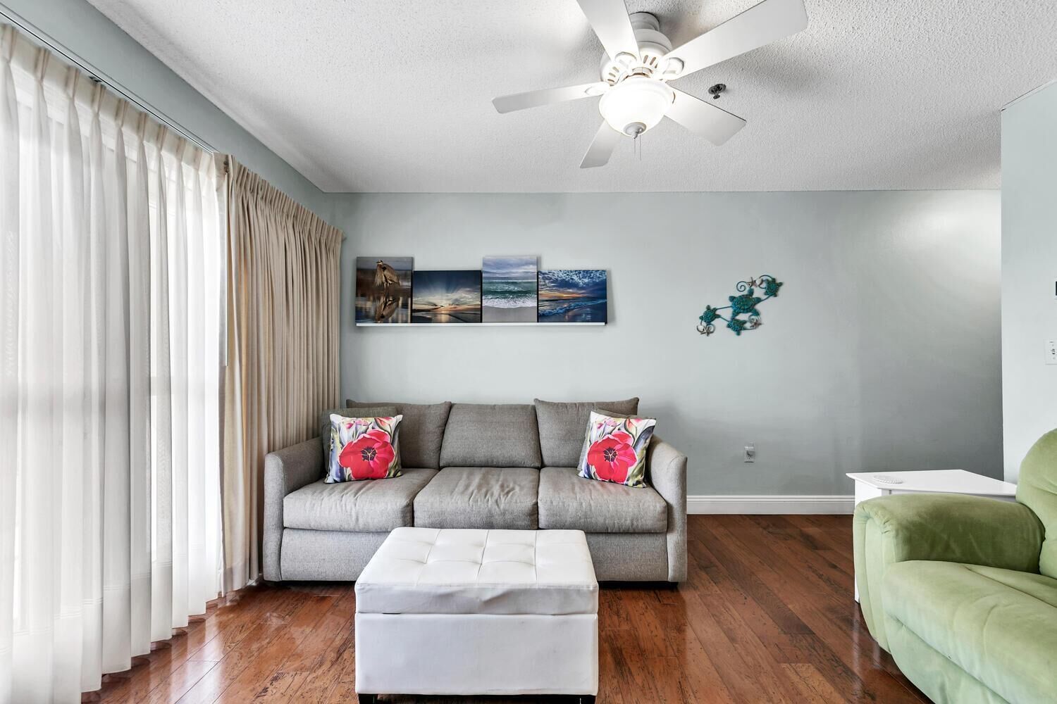 Un salon avec un canapé gris, un pouf blanc et un fauteuil vert. Trois œuvres d’art sont accrochées au mur et un ventilateur de plafond est installé.