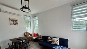 A living room with a dark blue sofa, a wooden dining table with chairs, a wall-mounted light fixture, and a ceiling fan.
