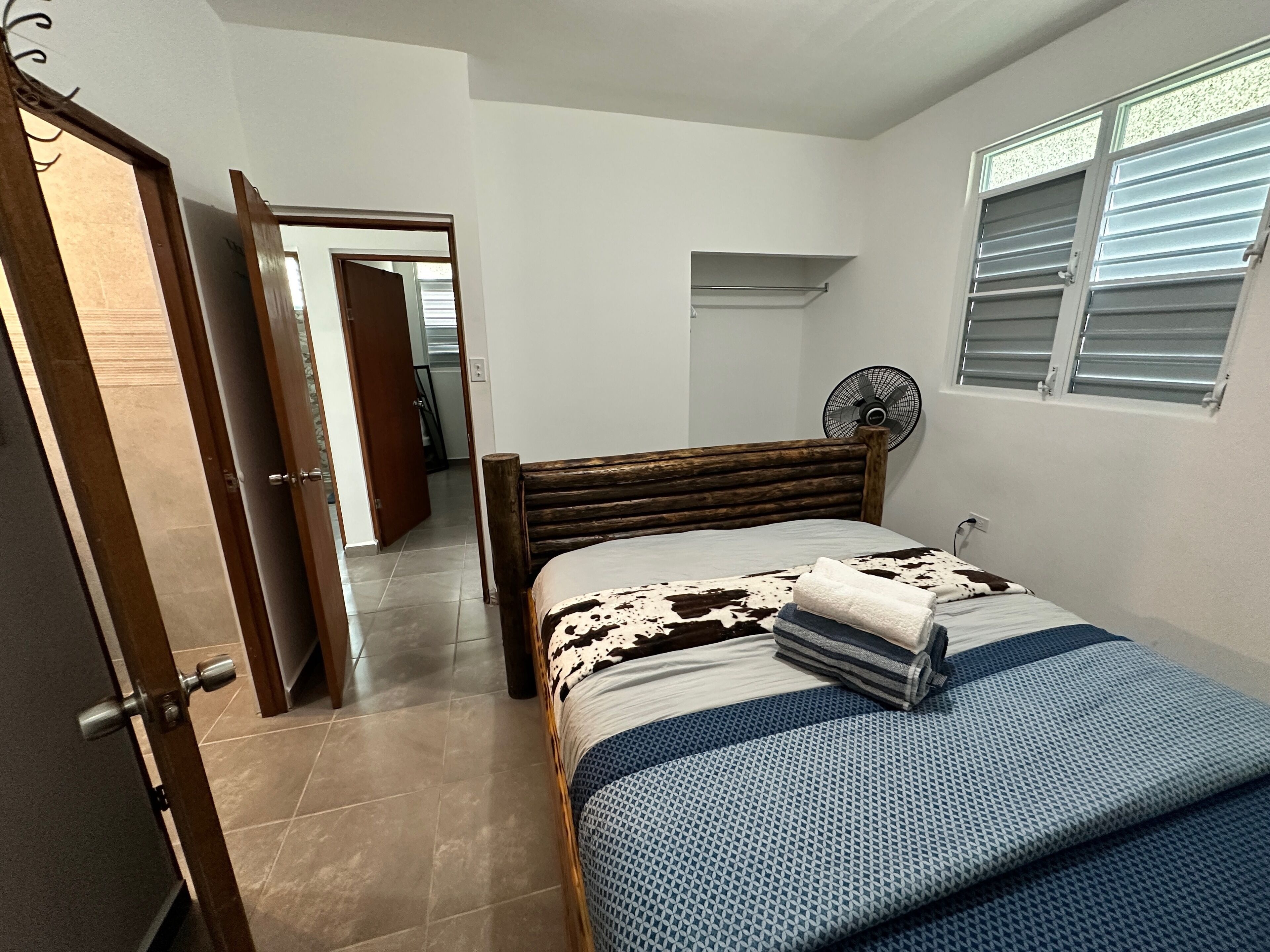 A bedroom with a wooden bed, a standing fan, and a window with blinds.
