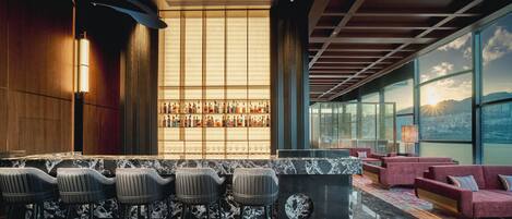 A modern bar area with a marble countertop, bar stools, and a large window offering a city view.